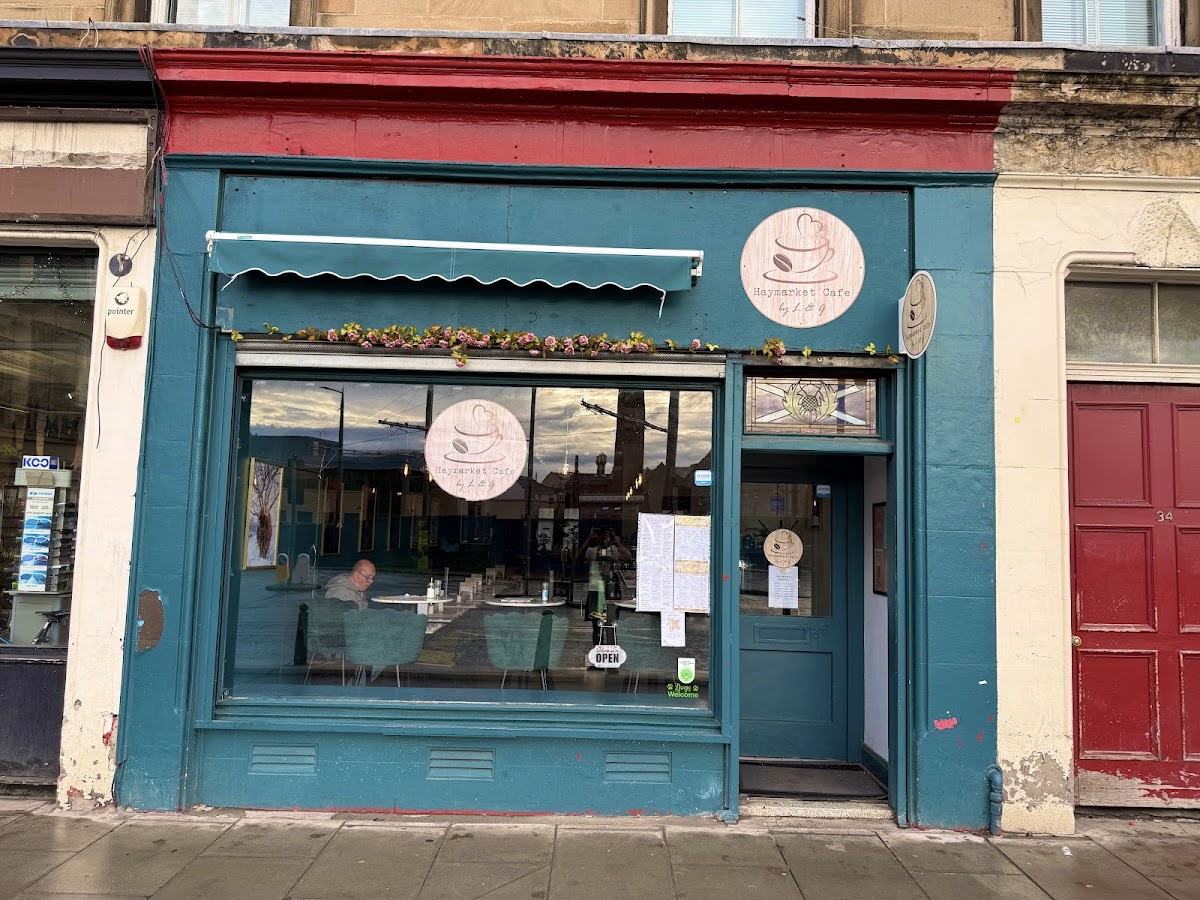 The teal shopfront of Haymarket Café by L&G with a red painted fascia, a round wooden sign showing a cup and a heart, and flowers along the window.