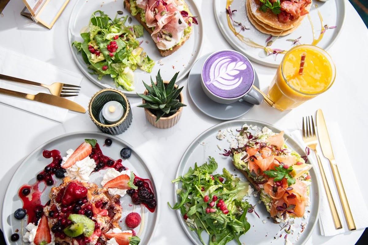 A table from above: avocado toast with prosciutto, a green salad, pancakes with strawberries, a berry waffle, a violet ube latte and a glass of orange juice.