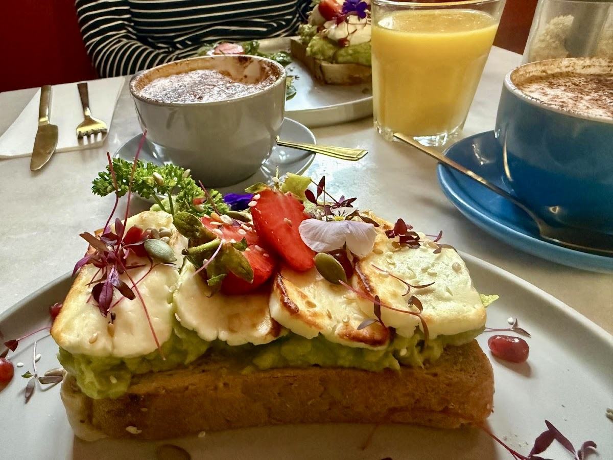 Avocado toast topped with grilled halloumi, strawberries, edible flowers and seeds, next to a dusted cappuccino.