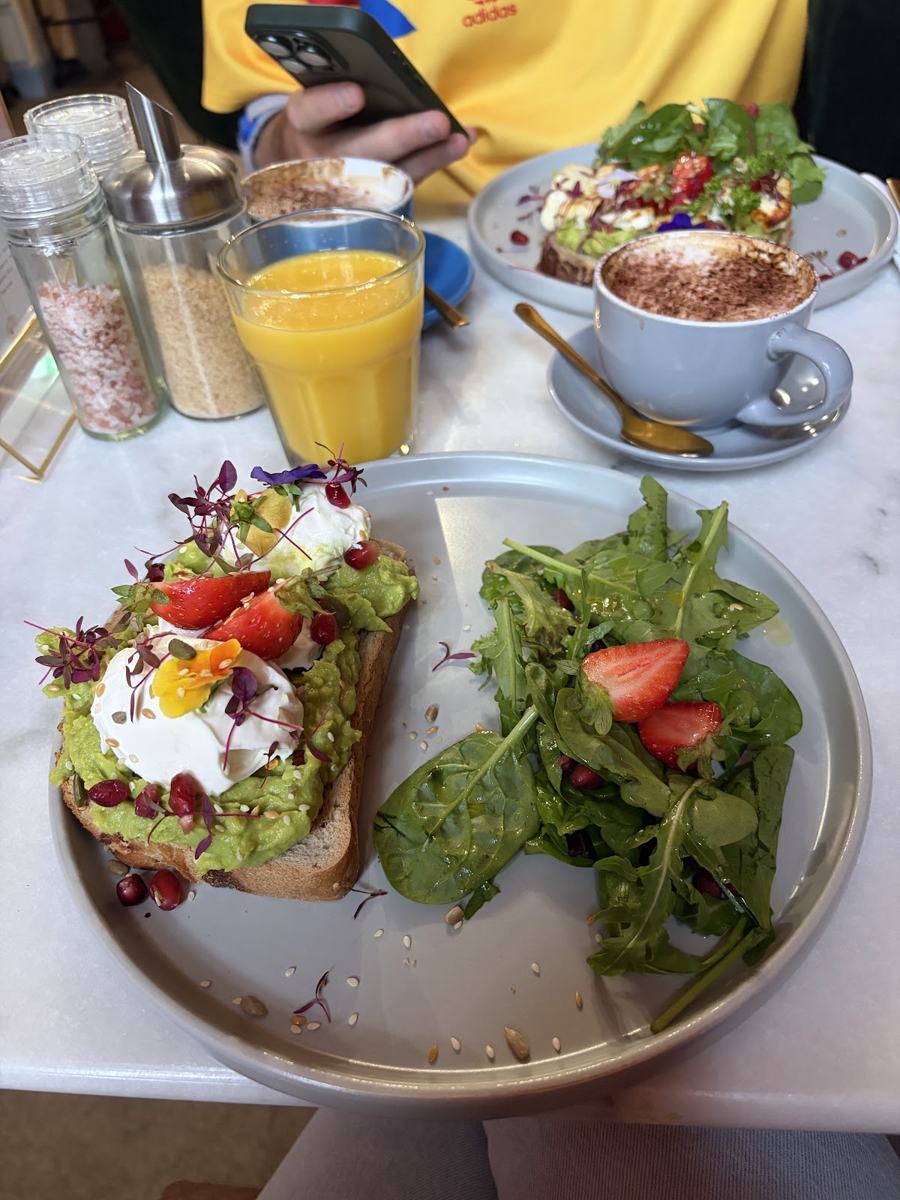 Avocado toast with two poached eggs and pomegranate seeds, served with a leafy salad, orange juice and a cappuccino on a marble table.