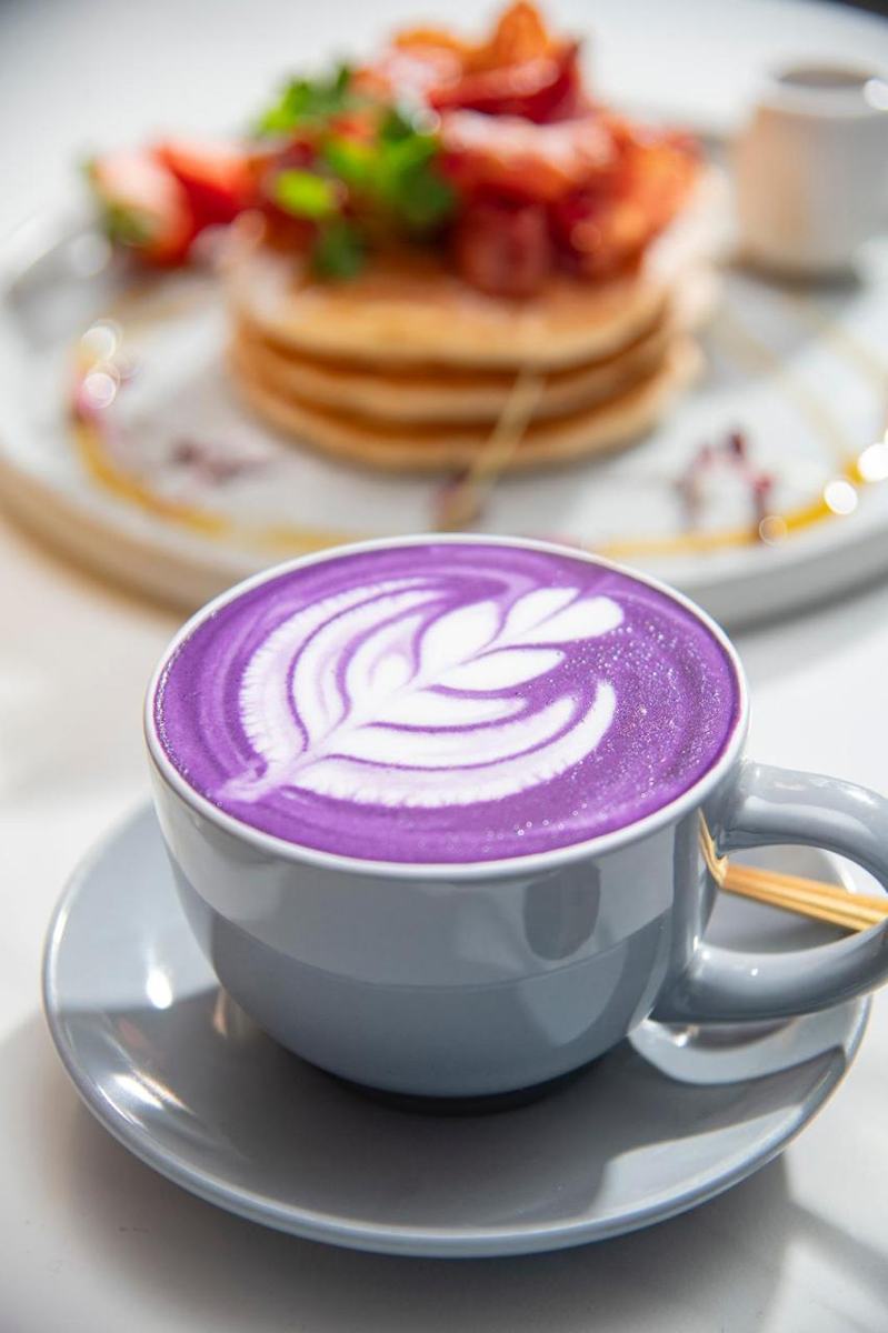 A violet ube latte with leaf art in a grey cup, a stack of pancakes with strawberries behind it.